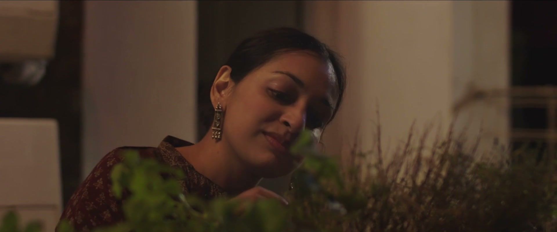 Mira, smiling and looking down, tends herbs in a balcony planter at dusk.
