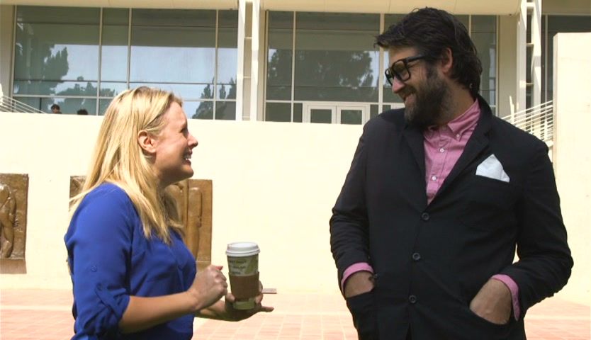 The presenter and a bearded man laughing in front of the art centre's glass facade.