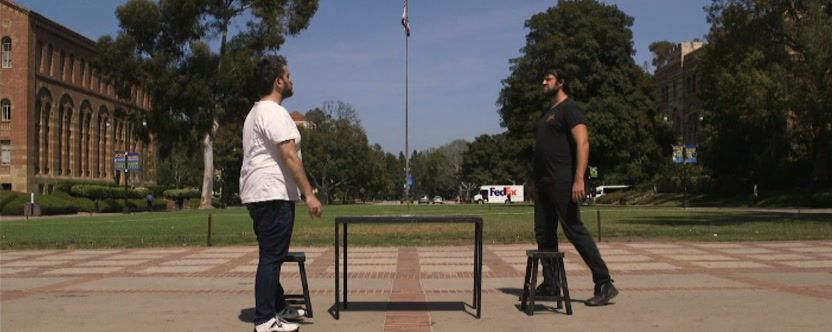 Two men face off, western-style, across a small table and two stools on a sunny campus plaza.