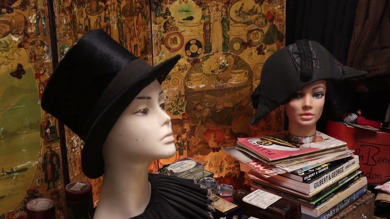 Two mannequin heads, one in a top hat and one in a bicorne, in front of a decoupage screen, with a stack of magazines and books. (opens a larger view)