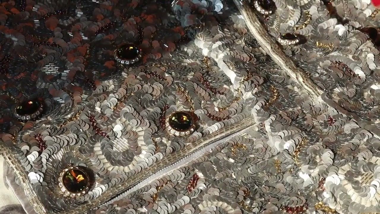 The texture of a silver sequin and bead garment with amber jewelled studs and a zip, filling the frame. (opens a larger view)