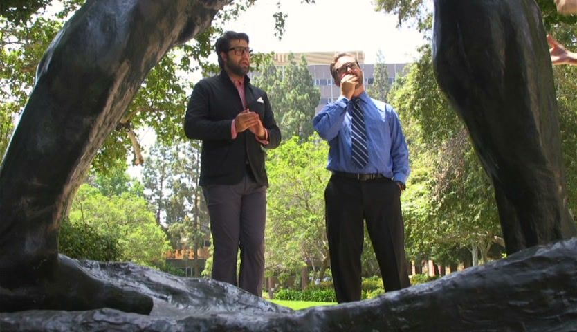Two men framed between the legs of a bronze striding figure in the sculpture garden, trees and a building behind.