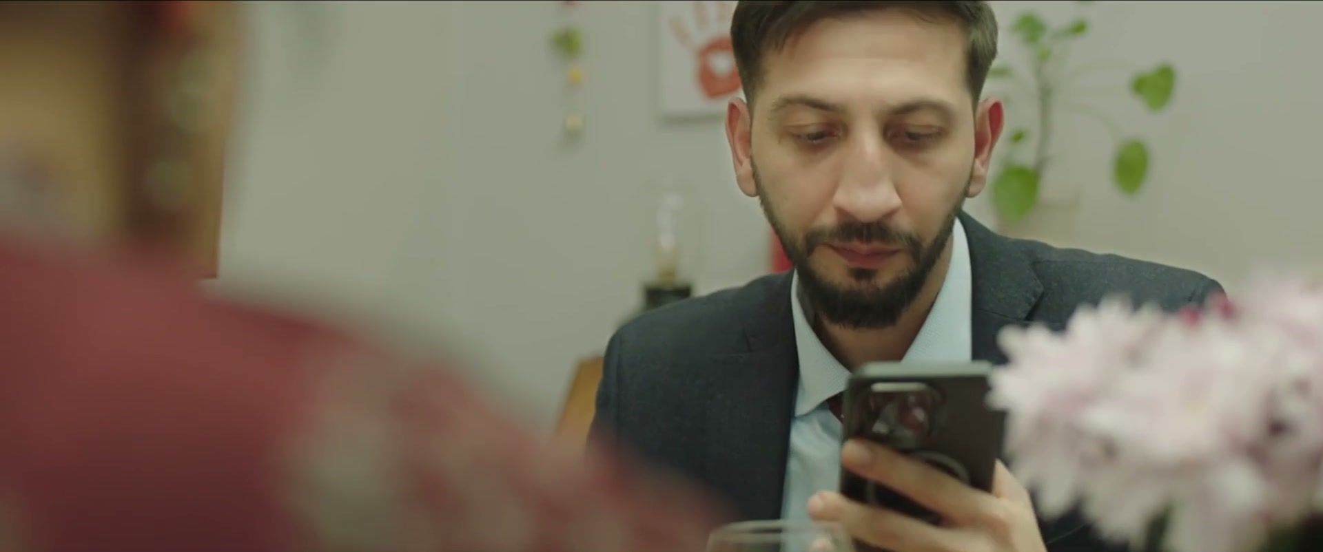 Ashwin looks at his phone at the table, seen over Mira's blurred shoulder, with soft pink flowers in the foreground.