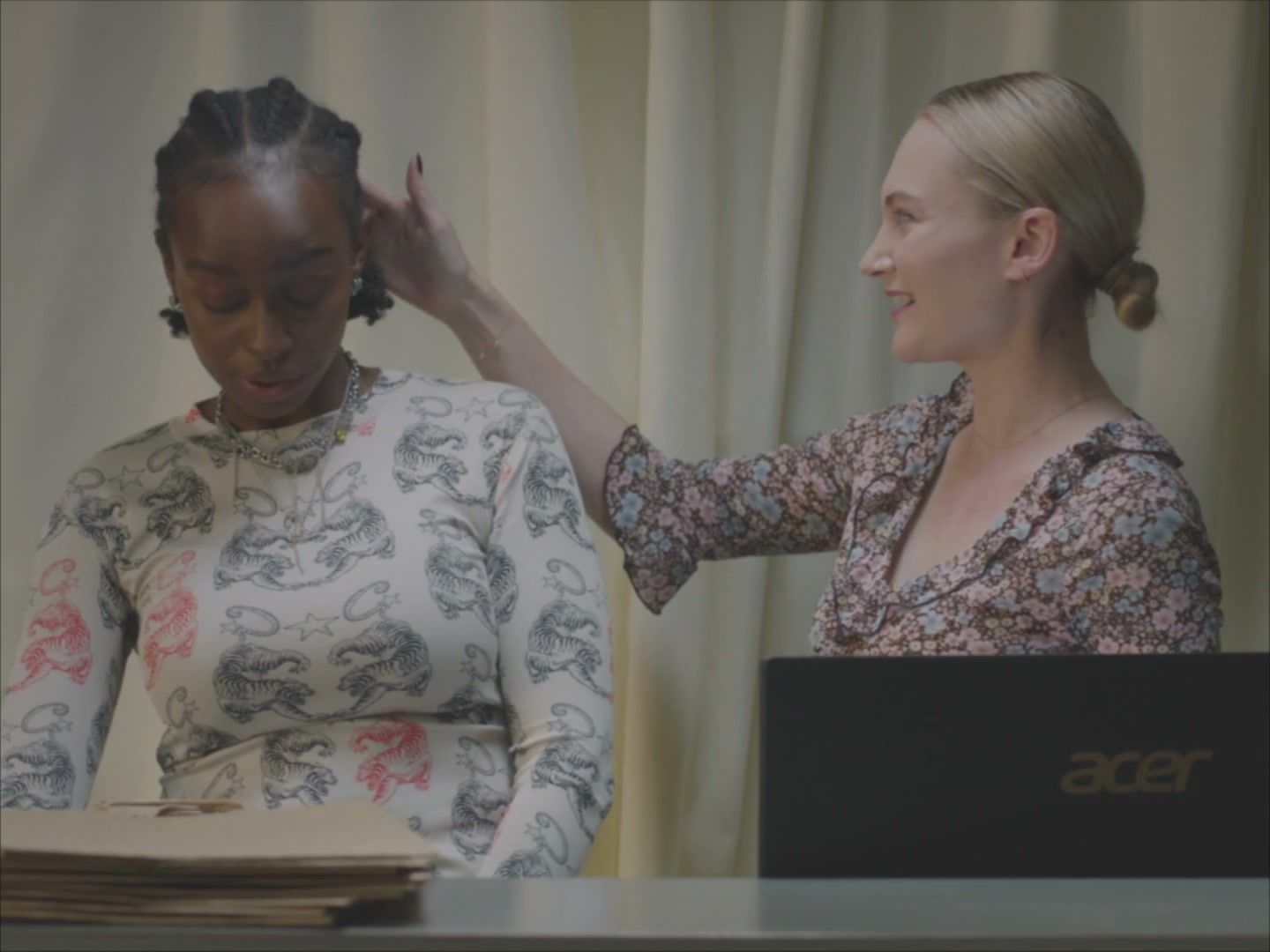 Two-shot at the desk: a blonde colleague reaches over to touch Lydia's braids while Lydia looks down. (opens a larger view)