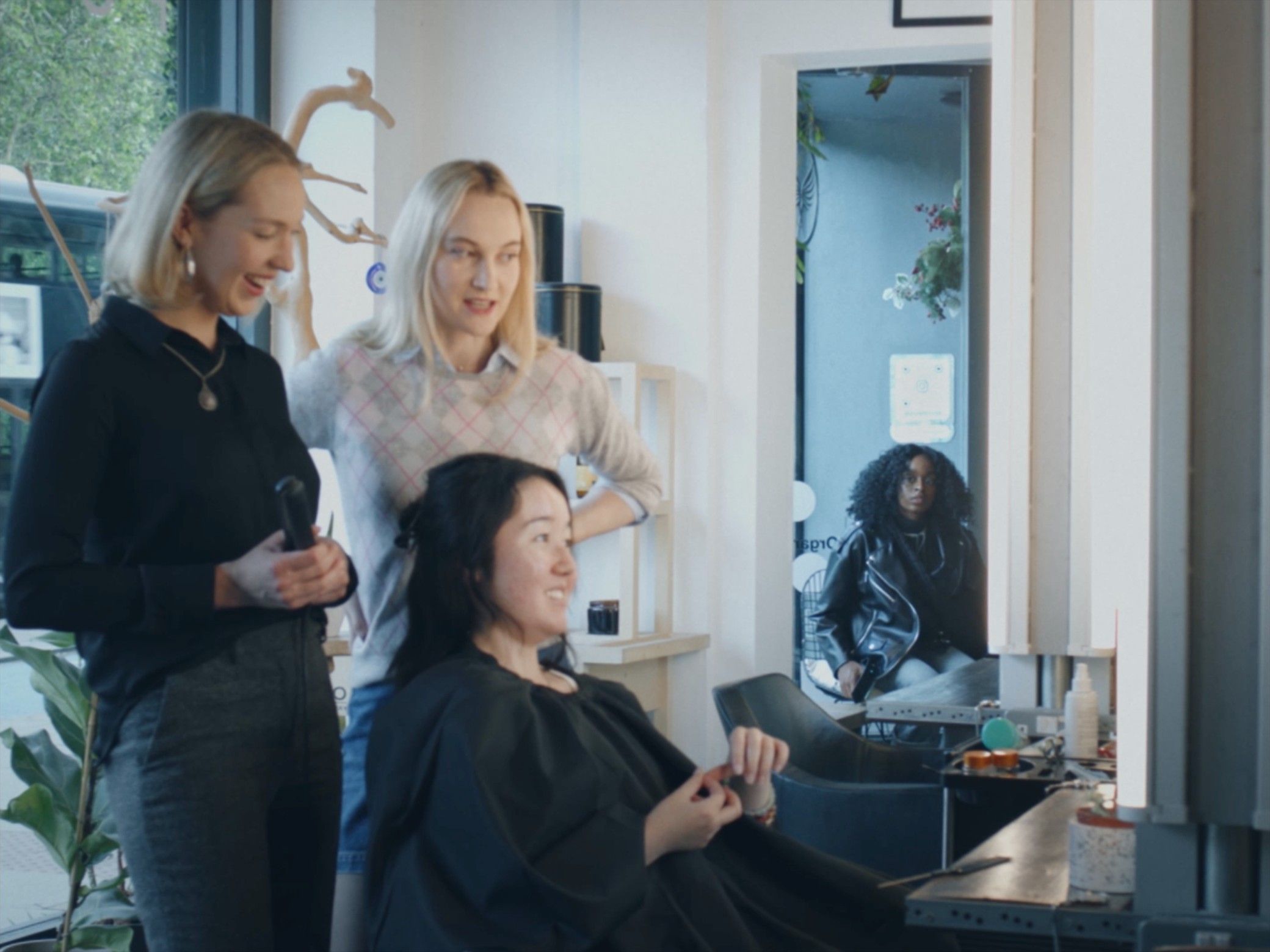 Two stylists stand over a seated client by the salon window; Lydia waits in the background, reflected in a mirror.