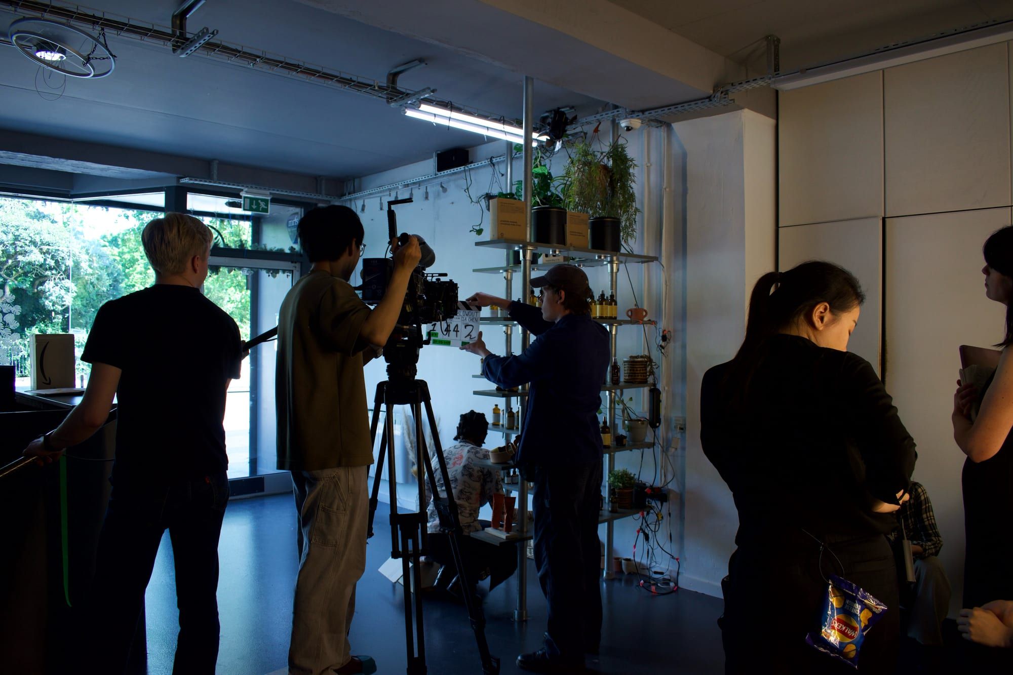 The crew in silhouette around the camera on its tripod in the salon, a bright window behind. (opens a larger view)