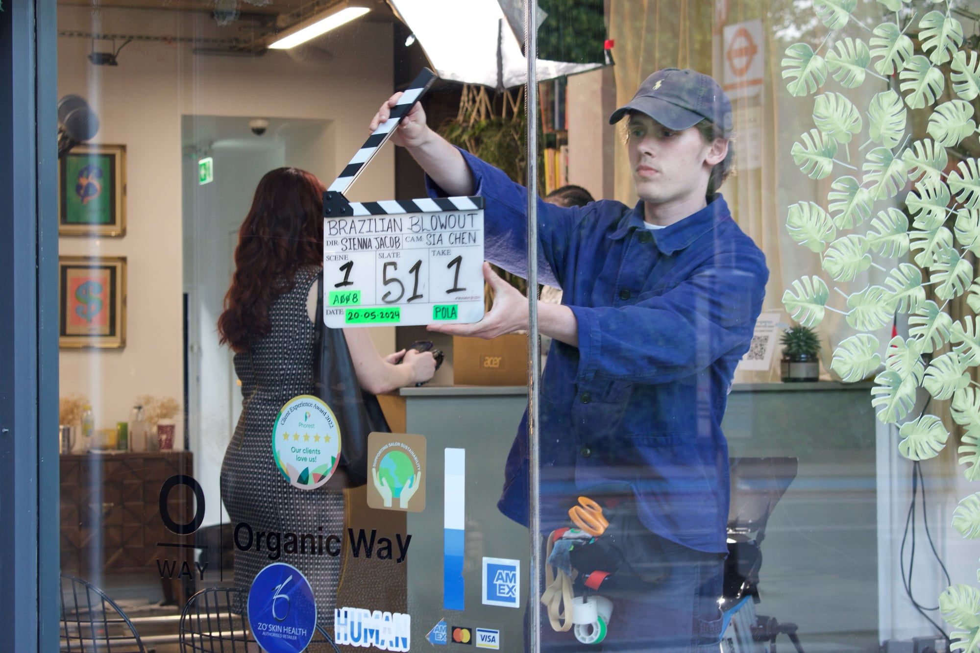 Through the salon window: a camera assistant holds up the slate (“Brazilian Blowout”, scene 51, take 1), actors behind.
